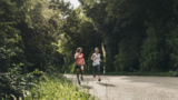 Zwei junge Frauen laufen bei sommerlichem Wetter auf einer Straße durch eine waldige Landschaft.